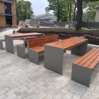 set 07 Outdoor plaza with long wooden benches on gray concrete bases, a paved area, and a construction zone behind a fence in the background.