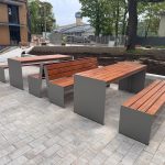 Outdoor plaza with long wooden benches on gray concrete bases, a paved area, and a construction zone behind a fence in the background.