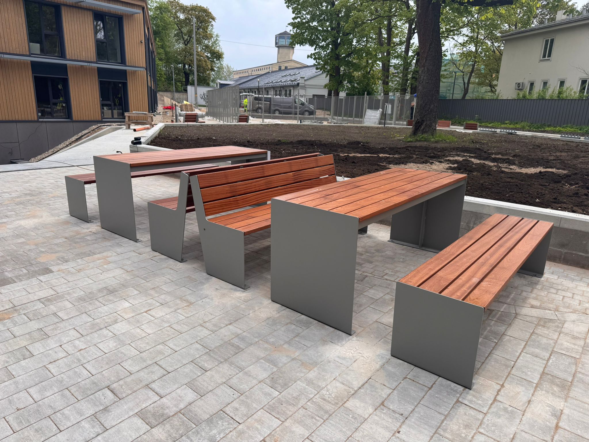 set 07 Outdoor plaza with long wooden benches on gray concrete bases, a paved area, and a construction zone behind a fence in the background.