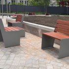 sols 600 Outdoor public seating: gray metal benches with wooden slats around a bare planting bed in a paved plaza, with a dark fence in the background and trees beyond.