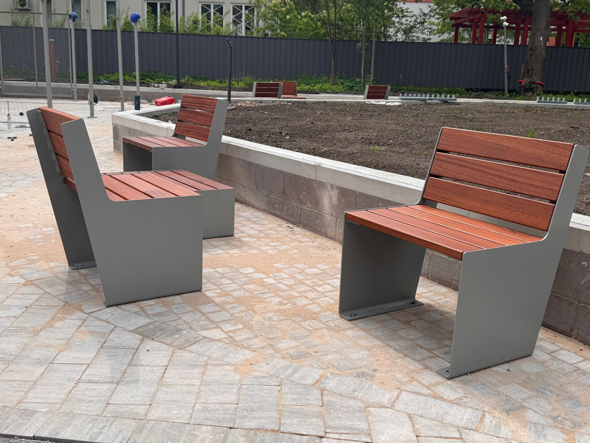 sols 600 Outdoor public seating: gray metal benches with wooden slats around a bare planting bed in a paved plaza, with a dark fence in the background and trees beyond.