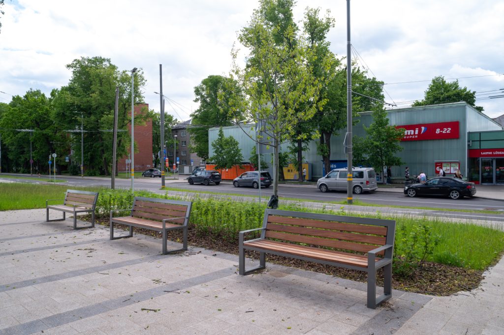 Three wooden benches with metal frames placed on a paved plaza, facing a busy street with cars and a storefront in the background.
