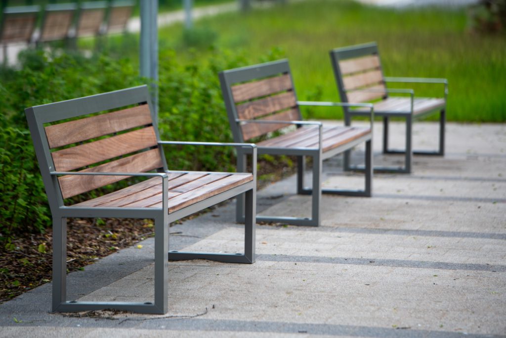 Three wooden slat benches with gray metal frames aligned along a paved walkway in a green park area.