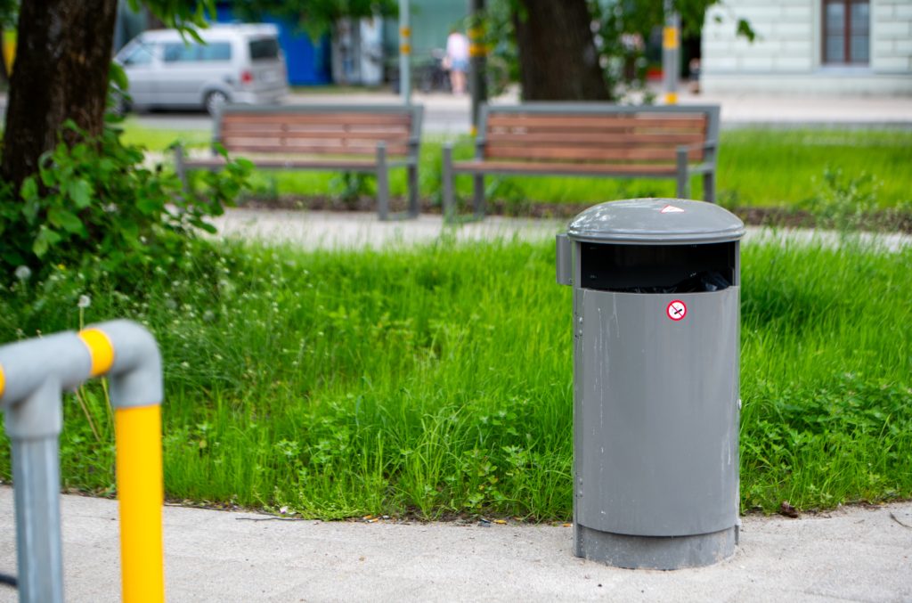 Grey metal outdoor trash can with open top and no-smoking sticker in a green park; blurred benches and a car in the background.