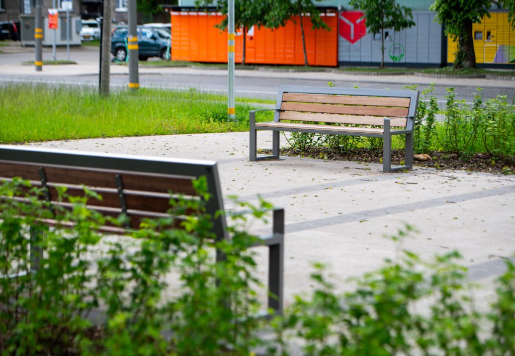 Empty wooden bench on a paved path with green grass nearby and bright orange storage containers in the background in an urban park setting.