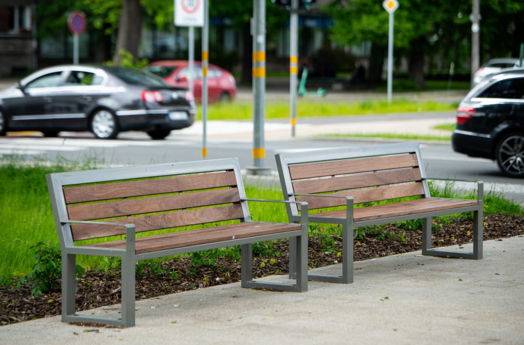 Two wooden benches with metal frames sit on a paved path beside a grassy area, with cars and street signs in the background.