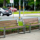 Two wooden benches with metal frames sit on a paved path beside a grassy area, with cars and street signs in the background.