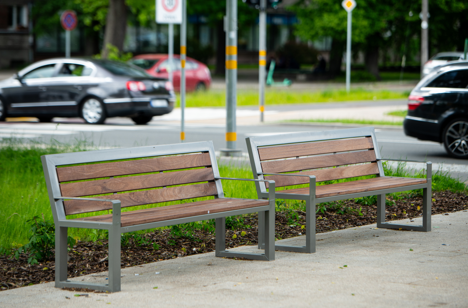 Two wooden benches with metal frames sit on a paved path beside a grassy area, with cars and street signs in the background.