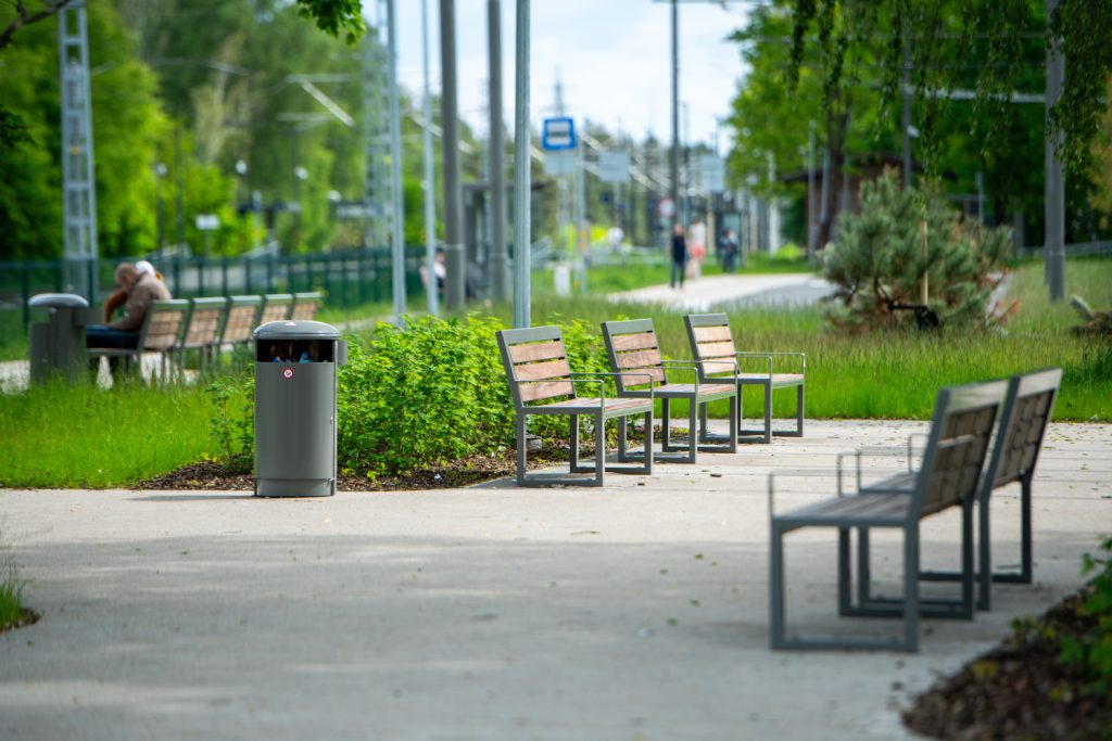 Row of wooden benches with metal frames along a paved park path, greenery on both sides and a trash can nearby.
